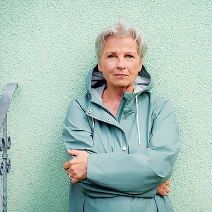 Portrait von fraujahnke mit cyan-farbener Jacke vor mint-grüner Wand, Arme verschränkt.