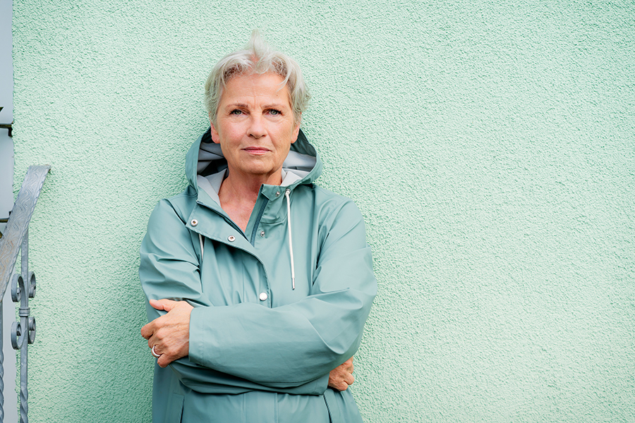 Portrait von fraujahnke mit cyan-farbener Jacke vor mint-grüner Wand, Arme verschränkt.